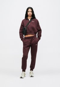 Bordeaux Nike huppari, jossa on vetoketju ja yhteensopivat joggerit, varustettuna joustavilla hihansuilla, lyhennetty muotoilu, yhdistettynä valkoisiin lenkkareihin ja pieneen mustaan laukkuun.