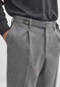 Pantalon en laine grise avec un devant plissé, doté de poches latérales et d'une fermeture par bouton à la taille. Texture douce visible.