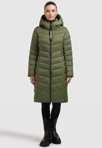 Zelena pernata jakna s kapuljačom, s patentnim zatvaračem sprijeda, prošivene teksture i dva bočna džepa. Nošena preko crnih helanki i masivnih crnih čizama.