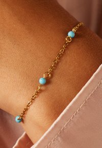 Gouden kettingarmband met drie lichtblauwe kralen. De armband heeft een delicate open schakelontwerp en een subtiele glans op een gebruinde pols.