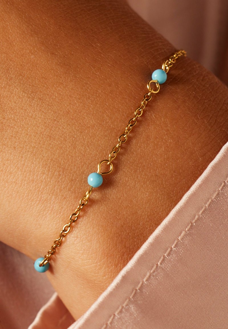 Gouden kettingarmband met drie lichtblauwe kralen. De armband heeft een delicate open schakelontwerp en een subtiele glans op een gebruinde pols.