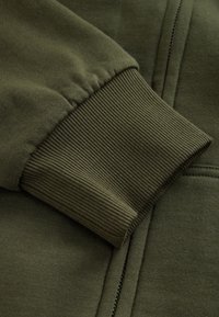 Olijfgroene trui-mouwboord met geribbelde textuur en elastisch ontwerp, met soepel stof dat zich uitstrekt naar een ritsopening.