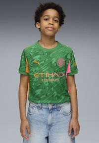 Maglietta verde a maniche corte con un motivo testurizzato, dettagli arancioni sulle spalle, grafiche rosa e logo del Manchester City sul petto. Pantaloni corti in denim.