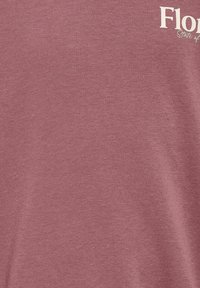 Κοντινή λήψη ενός μωβ υφάσματος με μέρος λευκού κειμένου που λέει "Flo" και "State of" στην πάνω δεξιά γωνία.