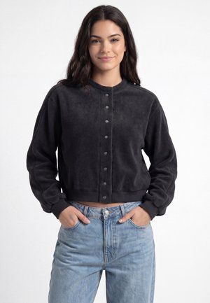 Ung kvinde stående med hænderne i lommerne på lyseblå jeans, iført en sort knaplukket langærmet jakke, der smiler let.