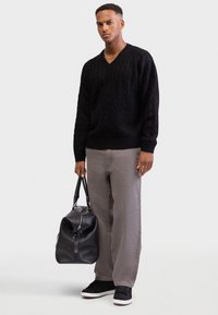 Czarny sweter w splot warkoczowy z dekoltem w serek, szare spodnie w luźnym fasonie, czarne tenisówki z białymi podeszwami oraz czarna torba podróżna z zamkiem błyskawicznym.