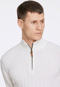 Weißer gestrickter Pullover mit hohem Kragen und Halb-Zip-Verschluss. Struktur mit Zopfmuster und einem Reißverschlusszipp aus braunem Leder.