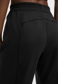 Nahaufnahme einer Person, die schwarze Sporthosen mit sichtbarer Naht und elastischem Bund trägt.