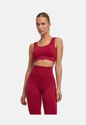 Mujer con sujetador deportivo rojo sin costuras y leggings de cintura alta, de pie contra un fondo claro liso con el pelo recogido.