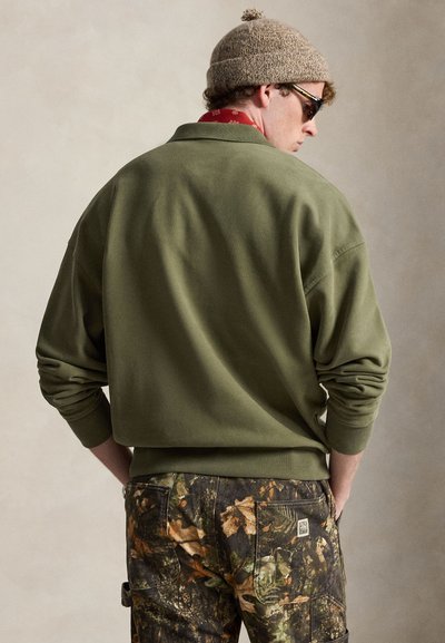 Άνδρας με ελιά πράσινο φούτερ, παντελόνι παραλλαγής, μπεζ πλεκτό σκουφί και γυαλιά ηλίου, όρθιος με τα χέρια στις τσέπες γυρισμένος προς τα πίσω.