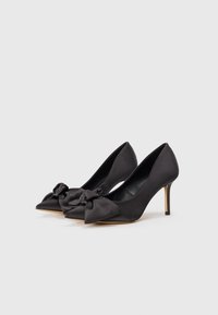 Pump in satin nero con tacco alto, punta affusolata, un fiocco decorativo sulla parte anteriore e un tacco sottile.