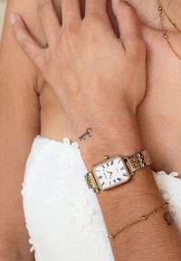 Montre en métal doré et argenté avec un cadran carré, un fond blanc, des chiffres romains noirs et un bracelet en acier inoxydable. Détails de tatouage complexes visibles.