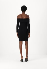 Abito nero a costine con spalle scoperte, maniche lunghe, silhouette aderente e dettaglio con bottoni sulla schiena. Abbinato a sandali neri con tacco a blocco.