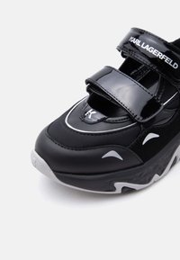 Μαύρο αθλητικό παπούτσι Karl Lagerfeld με παχιά λευκή σόλα πλατφόρμα, δύο λαμπερά λουράκια με βέλκρο και λευκές λογότυπες λεπτομέρειες στο επάνω μέρος και τη σόλα.