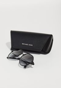 Occhiali da sole pilota neri con lenti scure, montatura in metallo e puntali in silicone, accanto a un astuccio coordinato in finto cuoio nero con logo.