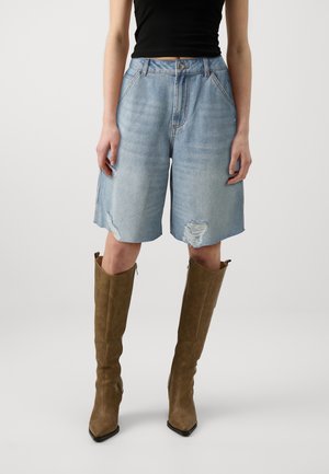 Person iført lyseblå slidte denimshorts, sort ærmeløs top og knæhøje brune støvler, stående mod en ensfarvet baggrund.