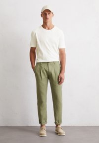 Hellbeige T-Shirt mit kurzen Ärmeln; olivgrüne Leinenhose mit Falten; beige Schuhe; Modell steht vor einem neutralen Hintergrund.