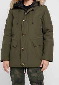 Parka de color verde oliva con capucha forrada de pelo sintético, botones expuestos y cordón ajustable. Cuenta con dos bolsillos frontales y puños ajustables.