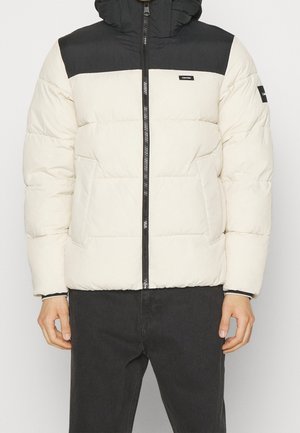Homme portant une doudoune blanche et noire avec une fermeture éclair à l'avant et deux poches latérales, associée à un pantalon noir.