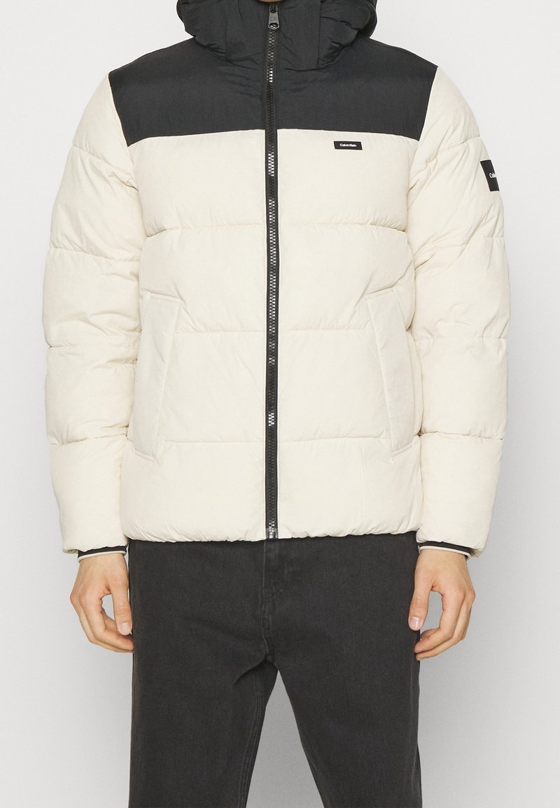 Homme portant une doudoune blanche et noire avec une fermeture éclair à l'avant et deux poches latérales, associée à un pantalon noir.