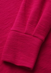 Top a maniche lunghe in maglia fucsia che mostra un tessuto testurizzato con polsini a coste. Il materiale è morbido e presenta sottili variazioni di tonalità.