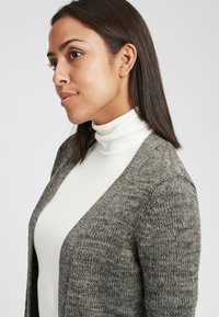 Gray knit cardigan with a open front design, textured fabric, and long sleeves, worn over a white turtleneck. Smooth and cozy appearance.