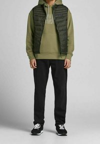 Gilet imbottito verde sopra un felpa oliva, abbinato a jeans neri e sneaker nere. Il gilet ha una chiusura con zip frontale e due tasche laterali.