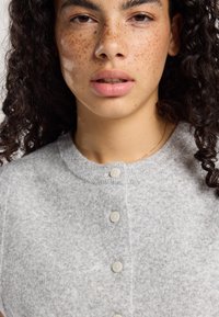 Gray knit cardigan with a round neck, featuring five light-colored buttons and a soft, textured fabric. Curly hair visible at the edges.