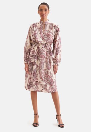 Femme debout portant une robe à manches longues, longueur genoux, avec des motifs cachemire rouges et roses et des sandales noires à talons hauts.