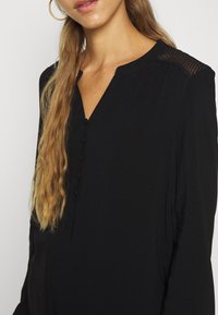 Blouse noire à manches longues avec col en V, patte de boutonnage et détails texturés aux épaules. Tissu doux et fluide avec une finition lisse.