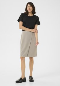 Zwarte t-shirt met korte mouwen gecombineerd met een beige wikkelrok tot de knie. Model draagt glanzende zwarte instappers. Effen achtergrond.