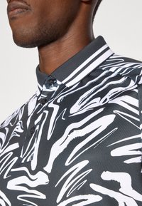 Polo preto com padrões abstratos brancos, apresentando um colarinho com duas riscas brancas e um pequeno logótipo no peito. Textura suave.