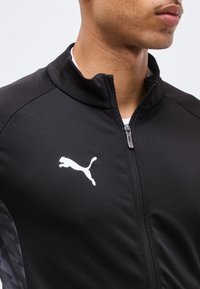 Άνδρας που φοράει μαύρο μπουφάν με φερμουάρ και λευκό λογότυπο Puma στο στήθος, εμφανιζόμενος από τον λαιμό μέχρι τη μέση, με απλό φόντο.