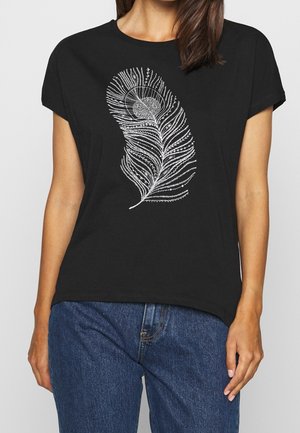 Kvinde iført en sort t-shirt med et detaljeret hvidt påfuglefjer-design, kombineret med blå denimjeans.