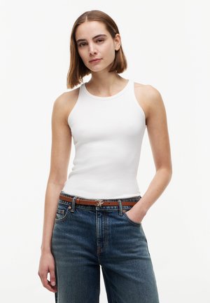 Jeune femme aux cheveux bruns lisses, portant un haut blanc sans manches, un jean bleu et une ceinture marron, debout avec une main dans la poche.