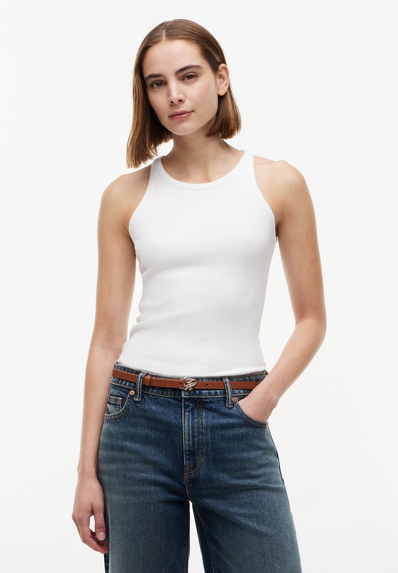 Νέα γυναίκα με ίσια καστανά μαλλιά, φορώντας λευκό αμάνικο τοπ, μπλε τζιν και καφέ ζώνη, στέκεται με το ένα χέρι στην τσέπη.