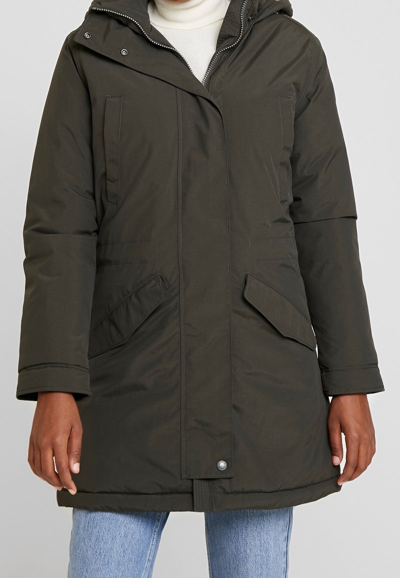 Donkergroene gewatteerde parka met klepzakken, voorrits, drukknoopsluiting en een aangesloten capuchon, gedragen over een witte coltrui en lichtblauwe jeans.