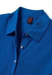 Polo blu realizzato in misto cotone. Presenta un colletto classico, bottoni frontali in azzurro chiaro e un'etichetta cucita con colori nero e rosso.