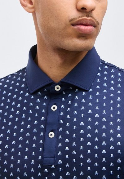 Άνδρας που φοράει μπλε ναυτικό πόλο πουκάμισο με λευκό μοτίβο κρανίου και σταυρωτών οστών και τρία λευκά κουμπιά στην πατιλέτα.