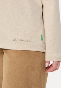 Béžová košeľa s dlhým rukávom, s jemnou textúrou, obsahujúca vyšitú VAUDE logo na leme a malú zelenú štítok na boku. Pod ňou sú nohavice z manšestru.