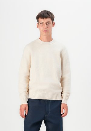 Un sweater beige clair en tissu doux et texturé avec un col rond et des poignets et un ourlet côtelés. Associé à un pantalon bleu marine foncé.