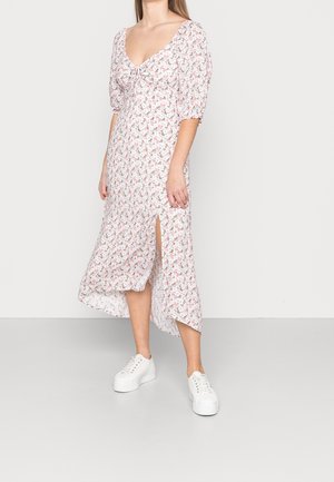 Frau trägt ein weißes Midikleid mit pinkem Blumendruck, Puffärmeln und seitlichem Schlitz, kombiniert mit weißen Plateau-Sneakern, steht auf grauem Boden.