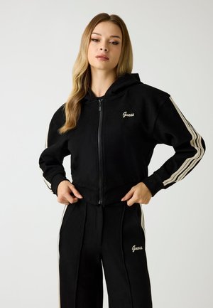 Sweatshirt noir zippé avec capuche, présentant des rayures crème le long des manches et le logo "Guess" brodé à l'avant, assorti à un pantalon noir.