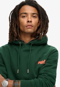 Felpa verde con cappuccio e lacci, con un logo arancione sul petto. Include una camicia a scacchi visibile ai polsini.