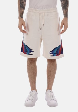 Mann trägt weiße Shorts mit Kordelzug und bunten blauen und roten Federdesigns an den Seiten, kombiniert mit weißen Sneakers und einem weißen Hemd.