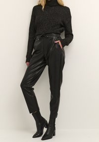 Pantalons en cuir noir à taille haute, avec poches avant et coupe relaxée, associés à un pull en maille noir à col montant et des bottines à talons noires.