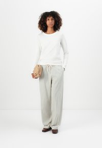 Femme aux cheveux bouclés portant un haut à manches longues blanc, un pantalon rayé ample, des chaussures foncées, tenant une pochette beige devant un fond blanc.