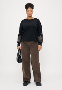 Μαύρο φούτερ με μοτίβο και διακοσμημένα μανίκια, συνδυασμένο με καφέ βελούδινες παντελόνες και μαύρα παπούτσια με πλατφόρμα. Κρατά μια μαύρη τσάντα.