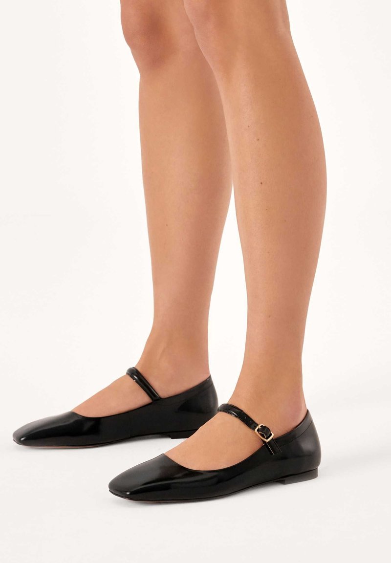 Jambes portant des ballerines Mary Jane noires brillantes avec de fines brides chevilles et une petite boucle sur fond blanc.
