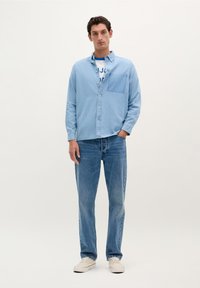 Camisa de mezclilla azul clara con un bolsillo en el pecho, combinada con jeans azul claros y zapatillas beige. El modelo está de pie frente a un fondo liso.
